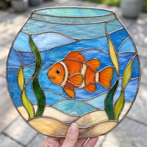 Può includere: Un pannello circolare in vetro colorato che raffigura un pesce pagliaccio che nuota in un acquario. Il pesce è arancione brillante con strisce bianche. Lo sfondo presenta acqua blu, alghe verdi e gialle e un fondale sabbioso, uniti dal metallo.
