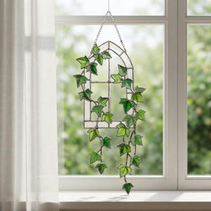 May include: A stained glass suncatcher featuring a clear arched window frame with trailing green ivy vines. The piece is constructed with soldered metal cames and hangs from a silver-toned chain, displaying various shades of green glass leaves.