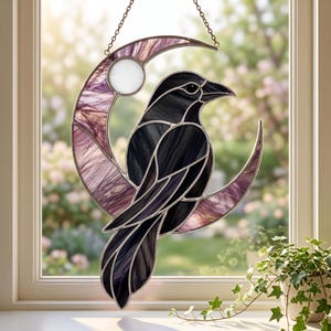 Könnte beinhalten: Ein Buntglas-Sonnenfänger mit einem schwarzen Vogel auf einer Mondsichel. Der Mond besteht aus durchscheinendem violettem Glas mit einem weißen kreisförmigen Akzent. Das Stück hängt an einer Metallkette und hat dunkle Metalllötstellen.