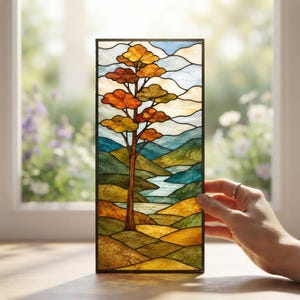 May include: A rectangular stained glass panel featuring a landscape scene with a central tree displaying orange and yellow foliage. The background depicts rolling green and blue hills, a winding river, and a pale sky, all framed by a dark border.