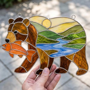 Può includere: Un acchiappasole in vetro colorato a forma di orso che tiene un pesce arancione. Il corpo dell'orso presenta un paesaggio con un sole giallo, colline verdi e un fiume blu. L'oggetto è realizzato con vetro marrone scuro e linee di saldatura.