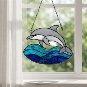 Könnte beinhalten: Ein Buntglas-Suncatcher mit einem grauen Delfin, der über stilisierte Meereswellen in Hellblau, Türkis und Dunkelblau springt. Das Stück ist mit dunklen Metall-Lötlinien eingefasst und hängt an einer schwarzen Metallkette.