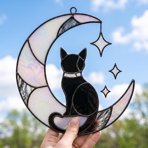 Op de afbeelding: Een glas-in-lood zonnevanger met een zwarte kat die op een halve maan van iriserend wit glas zit. Het ontwerp bevat drie vierpuntige steraccenten die aan de maan hangen, allemaal omlijnd met donkere metalen soldeerlijnen.
