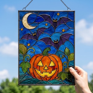 May include: A rectangular stained glass panel featuring a central orange jack-o'-lantern with a yellow face, surrounded by green leaves. The background depicts a dark blue night sky with a cream-colored crescent moon and three purple bats.
