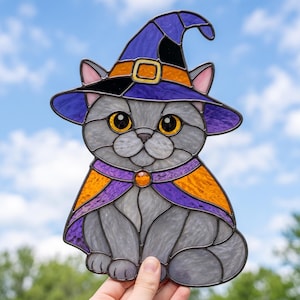 Può includere: Un acchiappasole in vetro colorato a forma di gatto grigio che indossa un cappello da strega viola con una fascia arancione e un mantello abbinato. Il gatto ha grandi occhi gialli e una piccola gemma arancione al centro del collare.