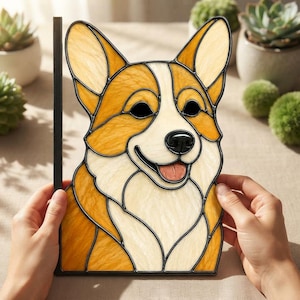Könnte beinhalten: Ein Buntglas-Sonnenfänger in Form eines Corgi-Hundekopfes mit bernsteinfarbenen, cremefarbenen und weißen Glasteilen, die mit schwarzem Metall umrandet sind. Der Hund hat eine schwarze Nase, dunkle Augen und ein offenes Maul mit einer rosa Zunge.