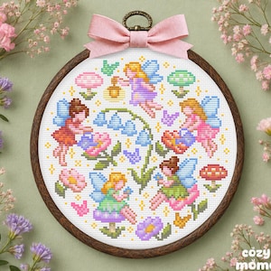 May include: A circular wooden embroidery hoop containing a cross-stitch pattern of four fairies with blue wings, colourful mushrooms, and blue bell-shaped flowers. A light pink fabric bow is attached to the top of the dark brown wooden frame.