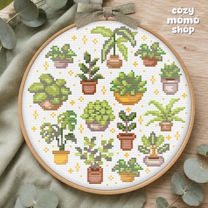 Pode incluir: Um bastidor de bordado circular de madeira contendo um padrão de ponto cruz de várias plantas em vasos em tons de verde, castanho e amarelo, acentuado com pequenas estrelas amarelas. O texto cozy momo shop está impresso no canto superior direito.