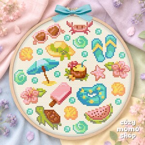 Pode incluir: Um bastidor de bordado circular de madeira contendo um padrão de ponto cruz com ícones de verão, incluindo óculos de sol cor-de-rosa, um caranguejo, chinelos, um guarda-sol, um coco, um gelado, uma tartaruga marinha e uma melancia.
