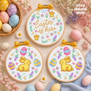 Pode incluir: Três bastidores de bordado circulares de madeira contendo padrões de ponto cruz. Um bastidor apresenta o texto "Easter is here" rodeado por flores e estrelas. Dois bastidores mostram um coelho amarelo rodeado por ovos coloridos, flores e estrelas.