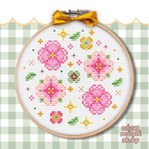 Pode incluir: Um bastidor de bordado em ponto cruz com um desenho floral de flores cor-de-rosa, folhas verdes e detalhes amarelos. O bordado está fixado num bastidor de madeira e amarrado com uma fita dourada. O texto "cozy momo shop" é visível.