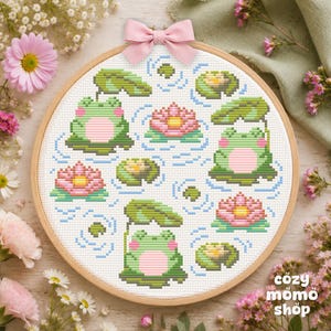 Pode incluir: Um bastidor de bordado circular de madeira contendo um padrão de ponto cruz de três sapos verdes com barrigas cor-de-rosa, flores de lótus cor-de-rosa e vitórias-régias sobre um fundo de tecido branco, com um pequeno laço de tecido rosa claro.