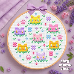 May include: A circular wooden embroidery hoop containing a cross-stitch pattern on white fabric. The design features six stylised cat faces in yellow, pink, and purple, interspersed with small flowers and paw prints. A light purple bow is attached.