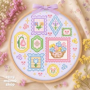 Pode incluir: Um bastidor de bordado em ponto cruz com uma variedade de desenhos florais em cores pastel. O design inclui flores emolduradas, uma cesta de flores e formas geométricas. Uma fita lavanda está amarrada no topo. O texto "cozy momo shop" está na parte inferior.