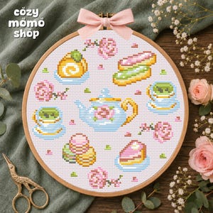 May include: A round wooden embroidery hoop containing a cross-stitch pattern of a light blue teapot, two teacups, and various pastries on a white fabric background. A pink ribbon is tied to the top of the hoop, which features floral accents.