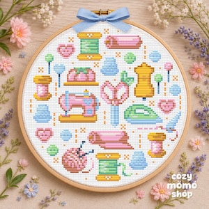 Pode incluir: Bastidor de bordado em ponto cruz com ícones de costura em cores pastel. Inclui uma máquina de costura rosa, tesouras, carretéis de linha e um manequim. Uma fita azul adorna o topo. O texto "cozy momo shop" está no canto inferior direito.
