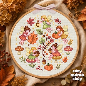 Pode incluir: Um bastidor de bordado circular de madeira contendo um padrão de ponto cruz em tecido branco. O design apresenta quatro fadas, folhas de outono, abóboras, cogumelos e bolotas em tons de laranja, vermelho, verde e castanho. Um laço rosa claro está atado no topo.