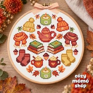 Pode incluir: Um bastidor de bordado circular contendo um padrão de ponto cruz com ícones de outono, incluindo um bule, livros empilhados, canecas com chantilly, gorros de malha, cachecóis e folhas, finalizado com um laço de fita cor de pêssego.