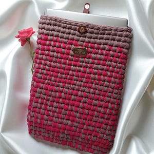 Puede incluir: Una funda de tableta hecha a mano con un patrón de ganchillo rosa y gris. La funda tiene un cierre de botón y una pequeña etiqueta dorada que dice "nuna". Está sobre una superficie de satén blanco.