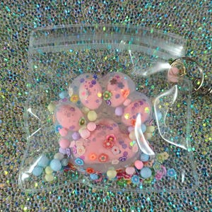 May include: A clear plastic bag containing a pink paw print-shaped charm with colorful embellishments. The charm is filled with glitter, small beads, and flower-shaped decorations. A silver-colored keychain is attached to the bag.
