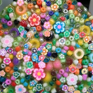 以下が含まれることがあります： 様々な色の小さな花とビーズが詰め込まれた、円形の樹脂アート作品。 花は主にピンク、オレンジ、青で、緑色の背景に配置されています。 この作品は、さまざまなビーズでいっぱいです。