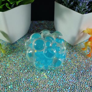 May include: A clear, bubble-shaped toy filled with blue liquid, surrounded by small plastic toy dragons in green and orange. The background features white planters with greenery and a glittery surface.