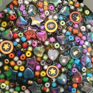 Puede incluir: Un diseño circular, estilo mosaico, con un fondo oscuro, lleno de cuentas de colores, estrellas, corazones y amuletos redondos. Los amuletos presentan símbolos de estrella, planeta, luna y ondas de radio. El diseño incorpora una variedad de colores, incluyendo rojo, naranja, amarillo, verde, azul y morado.