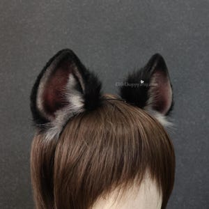 May include: A pair of black and gray faux fur wolf ears attached to a headband. The ears have a dark gray interior and are trimmed with white and gray fur. The headband is worn on a person with brown hair.