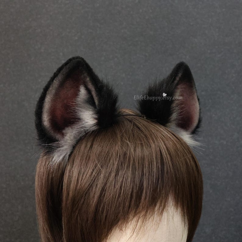May include: A pair of black and gray faux fur wolf ears attached to a headband. The ears have a dark gray interior and are trimmed with white and gray fur. The headband is worn on a person with brown hair.