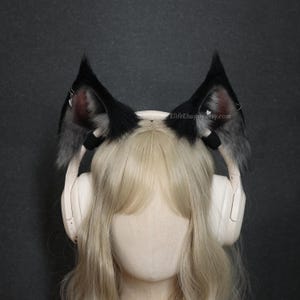 Puede incluir: Orejas de gato de piel sintética negra y gris con interior rosa, sujetas a una diadema. Las orejas se llevan sobre unos auriculares blancos. El texto "ElifeHappy.etsy.com" es visible en la oreja derecha.