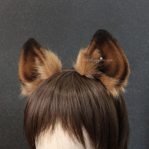 May include: A pair of brown and tan faux fur animal ears attached to a headband. The ears are upright and have a fluffy texture. The headband is thin and silver. The ears are worn on a person with brown hair.