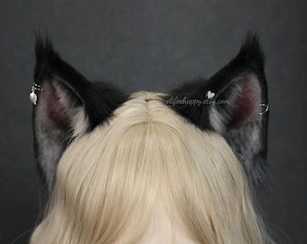 May include: Black and gray faux fur cat ears with a silver heart charm and hoop earrings. The ears are attached to a blonde wig. The text "elifeehappy.etsy.com" is visible on the right ear.