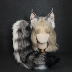Op de afbeelding: Een cosplay set met grijze en witte nepbont kattenoren, een bijpassende staart en een blonde pruik op een mannequin hoofd. De oren hebben roze binnendetails. De staart is overwegend grijs en wit met zwarte accenten.