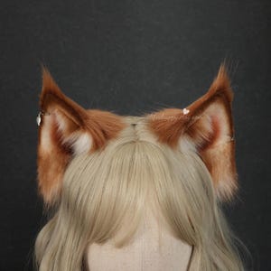 Op de afbeelding: Een paar vossenoren met een bruin en wit vachtontwerp. De oren zijn bevestigd aan een hoofdband en hebben kleine zilveren accenten. De oren worden gedragen door een persoon met blond haar.