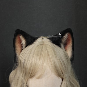 May include: Black and tan cat ears attached to a wig with long, light blonde hair. The ears are black on the outside with tan and pink interiors. The wig is styled with a center part. The background is a solid dark gray.