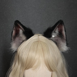 May include: A pair of faux fur animal ears in black and grey, featuring a gradient color pattern with darker tips and lighter inner sections. The ears are attached to a headband and are positioned atop a blonde wig with a center part.