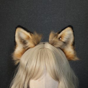 May include: A headband with a pair of faux fur fox ears. The ears are a blend of brown, tan, and black, with a fluffy texture. The ears are attached to a black headband. The background is a dark grey.
