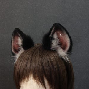 May include: A pair of black and gray faux fur cat ears. The ears are black on the outside and have a pinkish-gray interior. The ears are attached to a headband and are designed to be worn on the head. The text "Elifehappy.etsy.com" is visible on the right ear.