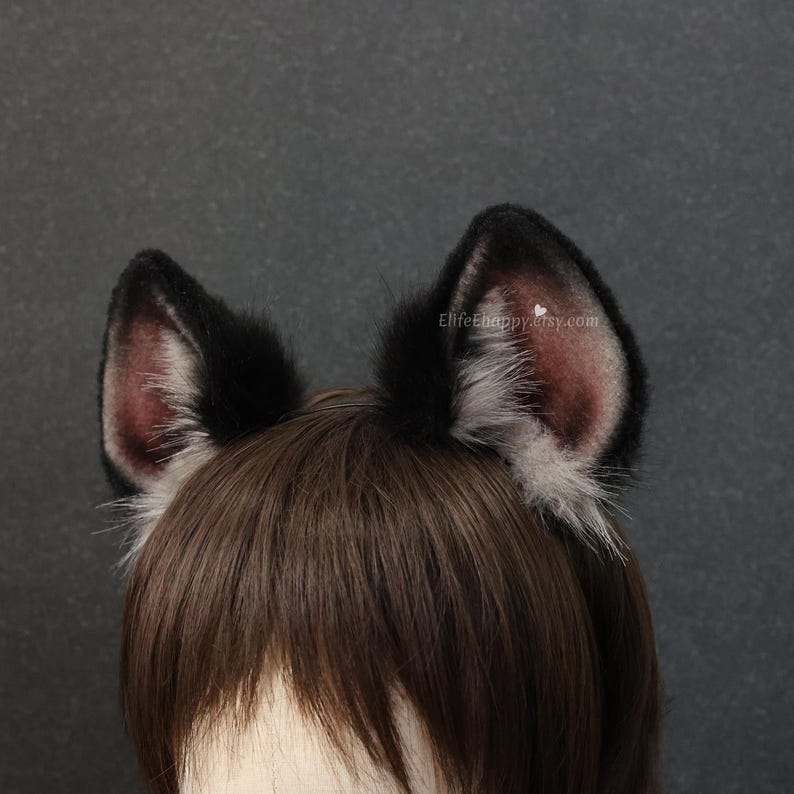 May include: A pair of black and gray faux fur cat ears. The ears are black on the outside and have a pinkish-gray interior. The ears are attached to a headband and are designed to be worn on the head. The text "Elifehappy.etsy.com" is visible on the right ear.