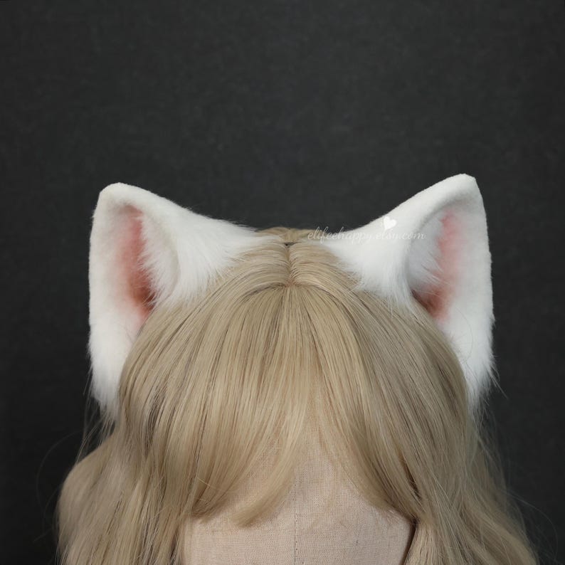 May include: A pair of white faux fur animal ears with soft pink inner detailing, attached to a headband worn over light blonde hair. The ears feature a plush texture and pointed shape, positioned symmetrically on top of the head.