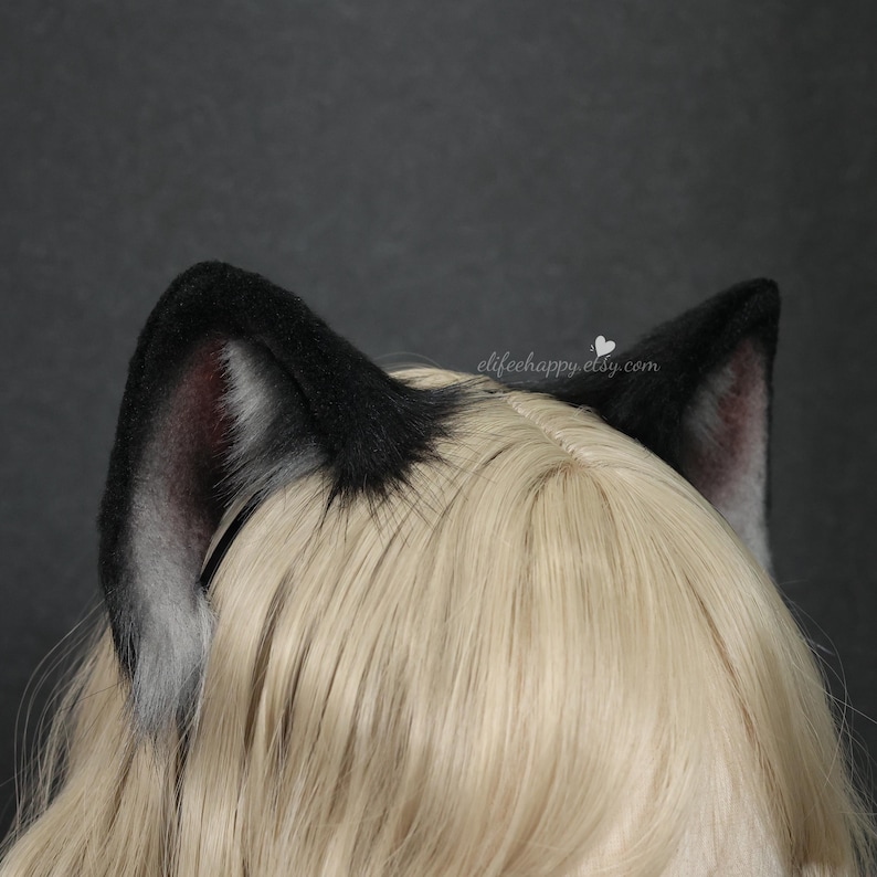 May include: A pair of black faux fur animal ears attached to a headband, positioned on top of straight blonde hair. The ears feature a dark black outer edge with a soft, light grey and white inner fur texture. The text elifehappy.etsy.com is visible.
