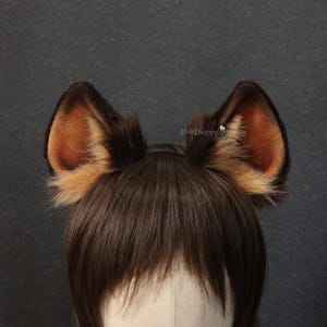 May include: A pair of brown and tan faux fur animal ears attached to a black headband. The ears have a realistic design with a darker brown outer edge and a lighter tan inner ear. The ears are set against a dark gray background.