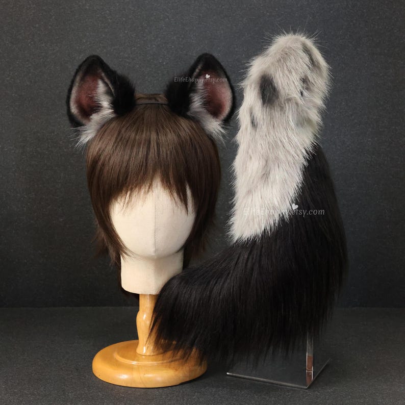 May include: A set of black and gray faux fur animal ears and tail. The ears are black with gray trim and pink inner ears. The tail is black with a gray and white section at the top. The ears are attached to a brown wig on a mannequin head.