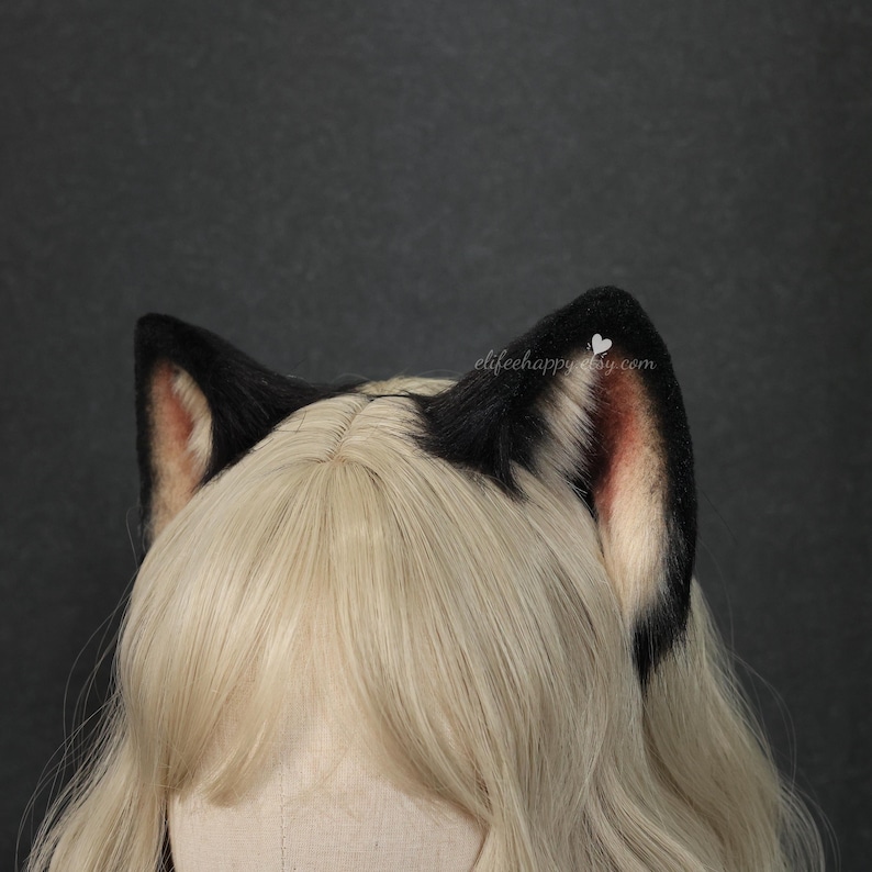 May include: A pair of black faux fur animal ears with pinkish-tan inner detailing, positioned atop a blonde wig. The text elifeehappy.etsy.com is visible in small white lettering near the right ear. The ears feature a soft, plush texture.