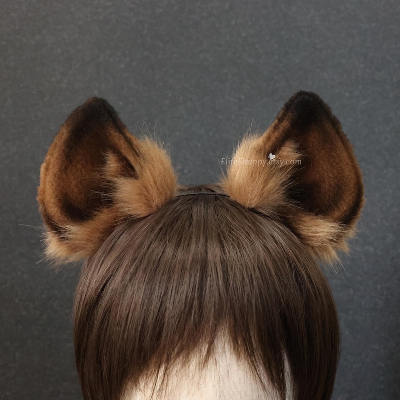 May include: A pair of brown and tan faux fur animal ears attached to a black headband. The ears are upright and have a fluffy texture, with darker brown tips. The background is a solid dark gray.