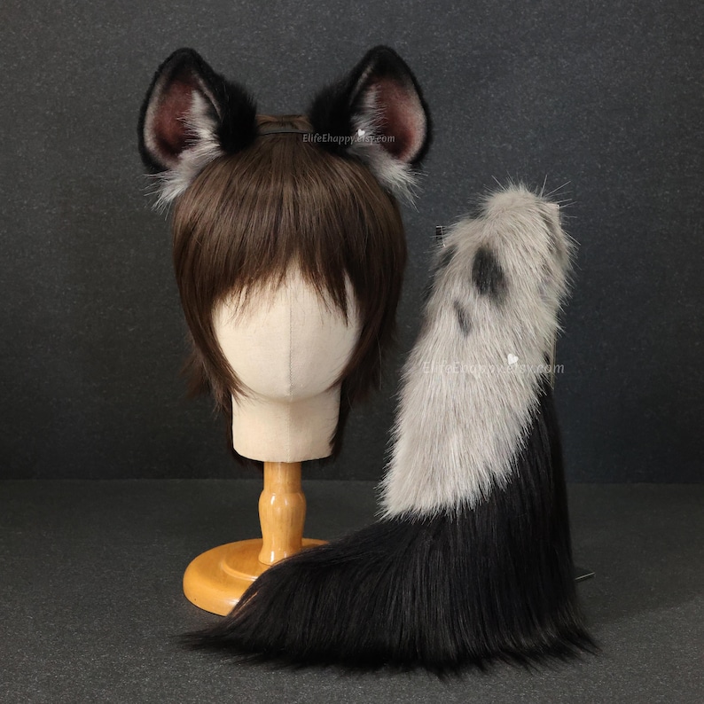 May include: A black and gray faux fur tail and matching ear headband set. The ears are black with pink and gray inner details. The tail is black with a gray and black spotted pattern. The set is displayed on a mannequin head.