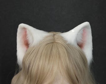 May include: White faux fur cat ears with pink inner ear detail. The ears are attached to a headband and are worn on the head. The ears are fluffy and stand upright. The background is a solid dark color.