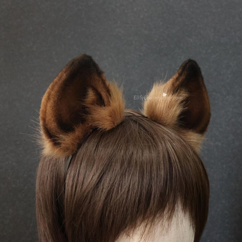 May include: A pair of brown and tan faux fur animal ears attached to a black headband. The ears are a darker brown at the tips and lighter brown at the base, with a fluffy texture. The ears are set against brown hair.