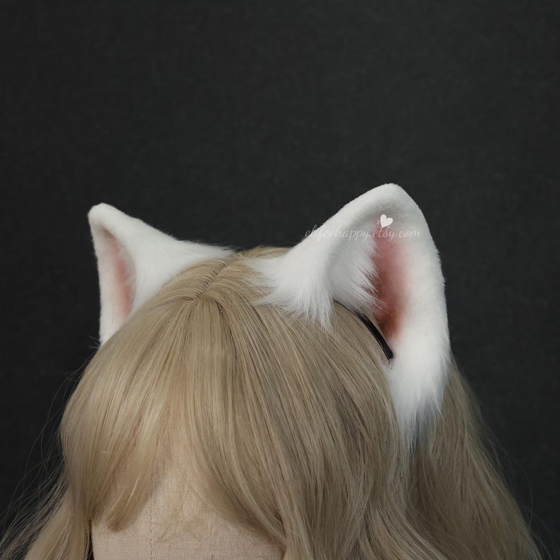 May include: A headband featuring two pointed, white faux fur animal ears with soft pink inner detailing. The ears are attached to a thin black band and are positioned atop a light blonde wig, showcasing a fluffy texture and a realistic shape.