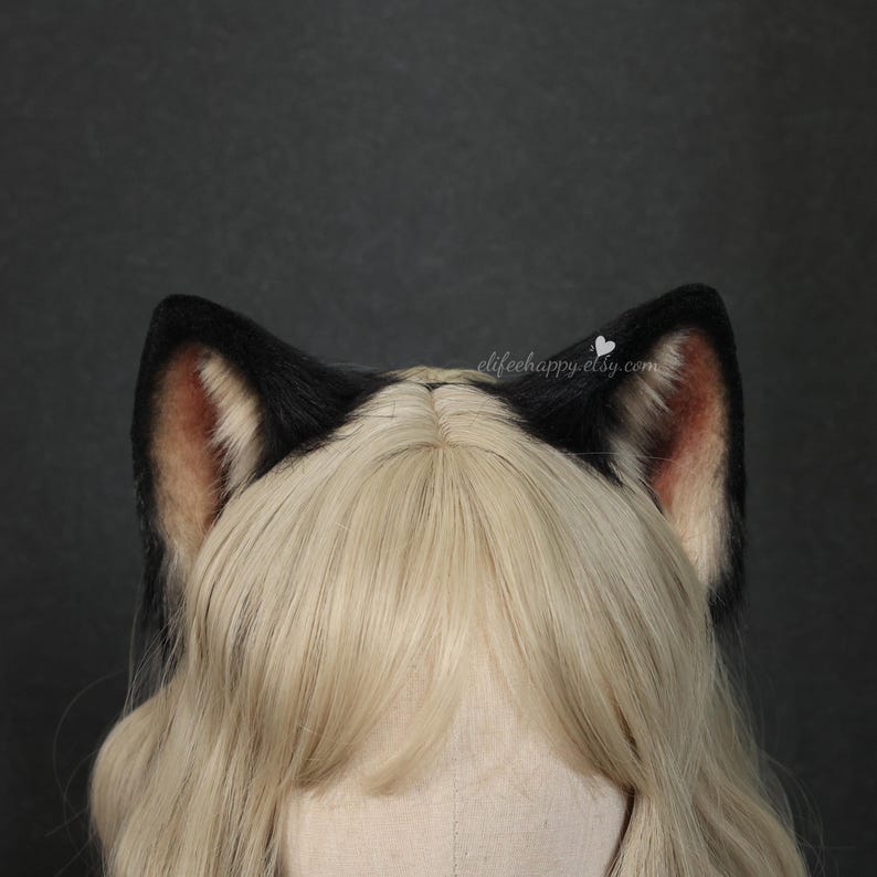 May include: A pair of black and white faux fur animal ears attached to a headband, positioned on a blonde wig. The inner ear features a soft pinkish hue, while the outer edges are dark black. The text elifeehappy.etsy.com is visible in white.